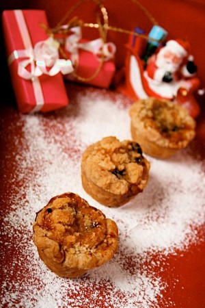 Mini Almond Crumble Mince Pies: on a red background with dusting of icing sugar like snow. In soft focus are dinky presents and a santa ornament.