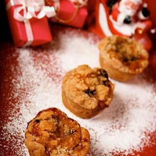 Mini Almond Crumble Mince Pies: on a red background with dusting of icing sugar like snow. In soft focus are dinky presents and a santa ornament.