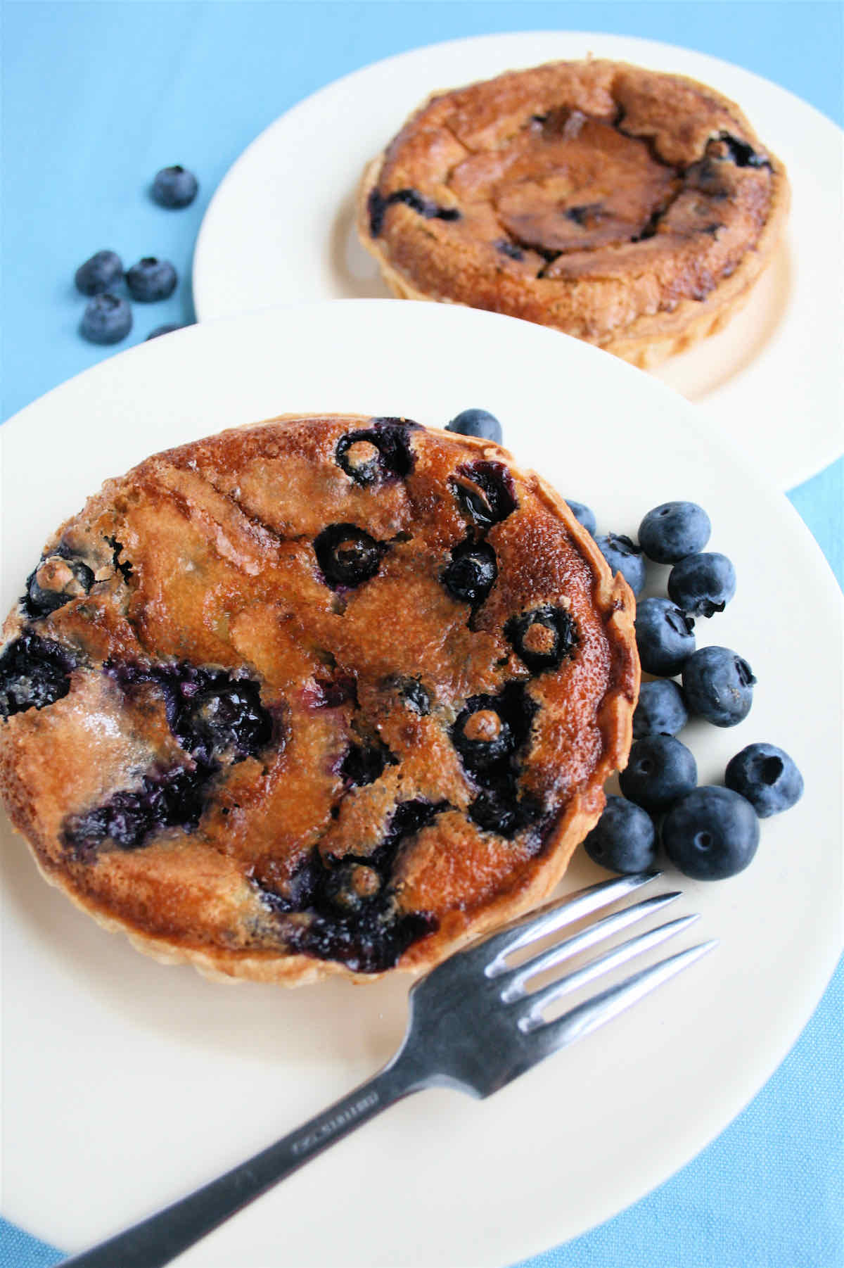 Blueberry Cinnamon Tarts: A golden blueberry tart on a white plate with raw blueberries and a fork.