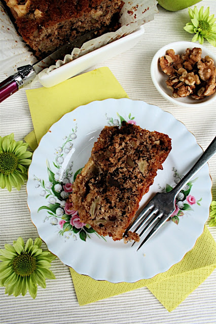 Apple Loaf Cake with Nuts and Chocolate: from above, a white plate with dessert fork, rectangle slice of loaf cake, on a surface with green flowers and paper napkin.