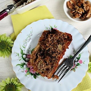 Apple Loaf Cake with Nuts and Chocolate: from above, a white plate with dessert fork, rectangle slice of loaf cake, on a surface with green flowers and paper napkin.