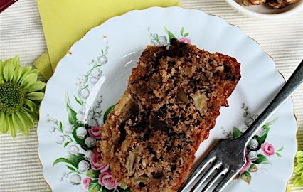 Apple Loaf Cake with Nuts and Chocolate: from above, a white plate with dessert fork, rectangle slice of loaf cake, on a surface with green flowers and paper napkin.