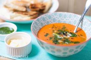 Roast tomato and red pepper soup topped with basil oil and shaved grana padano cheese served in pattern light blue bowls, smaller white dishes of the toppings are to the side.