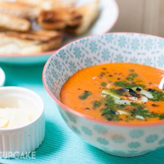 Roast tomato and red pepper soup topped with basil oil and shaved grana padano cheese served in pattern light blue bowls, smaller white dishes of the toppings are to the side.