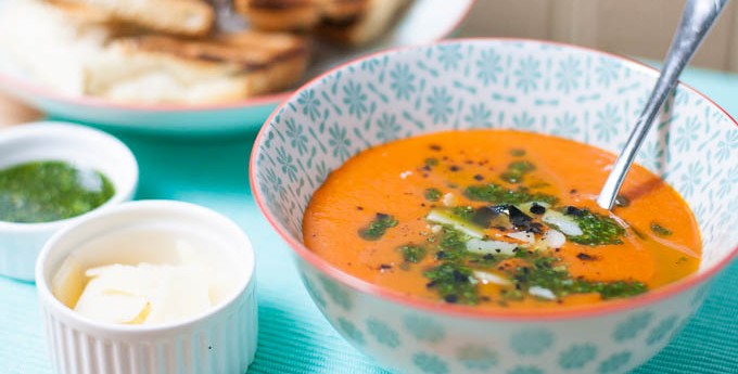 Roast tomato and red pepper soup topped with basil oil and shaved grana padano cheese served in pattern light blue bowls, smaller white dishes of the toppings are to the side.