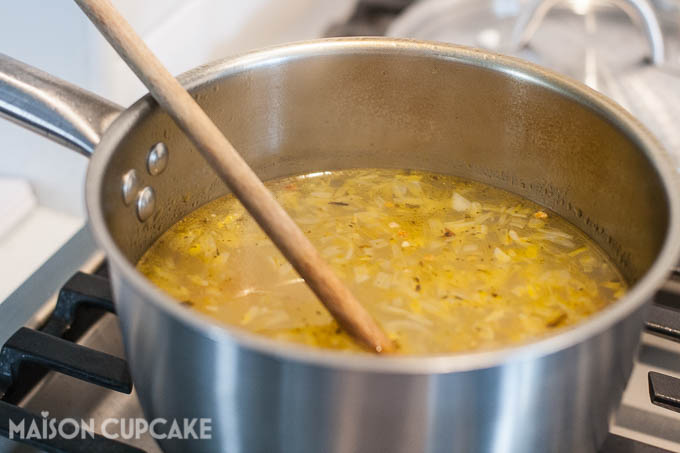 Stainless steel pan on a gas hob, filled with yellow liquid comprising vegetable stock and pieces of chopped vegetables. A wooden spoon is sticking out of the pan.