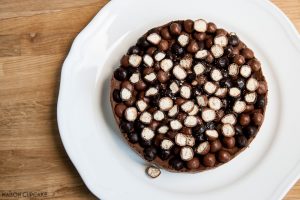 Chocolate Quark Cheesecake - Easy No Bake Recipe. Seen from above, chocolate cheesecake wtih crunchy biscuit pieces. A slice has been cut away and sites on a stripey blue plate with a fork. A bag of McVities Nibbles to the side.
