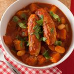 A white bowl of low fat sausage casserole with beans in tomato sauce upon a red gingham napkin with a fork.