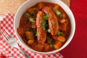 A white bowl of low fat sausage casserole with beans in tomato sauce upon a red gingham napkin with a fork.