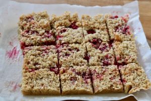 Raspberry flapjacks with coconut traybake, homemade and sliced into a dozen squares on baking paper. Fresh raspberry fruit is visible and has stained the paper.