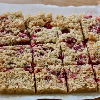 Raspberry flapjacks with coconut traybake, homemade and sliced into a dozen squares on baking paper. Fresh raspberry fruit is visible and has stained the paper.