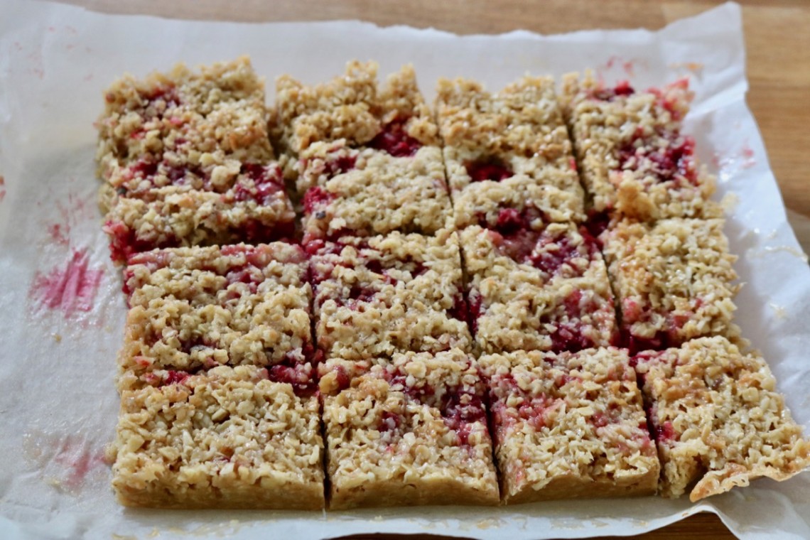 Raspberry flapjacks with coconut traybake, homemade and sliced into a dozen squares on baking paper. Fresh raspberry fruit is visible and has stained the paper.