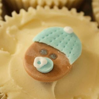 Baby shower cupcake decorated with cream buttercream icing and a sugarpaste baby face with bobble hat and dummy.