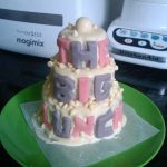 A small tiered cake covered in white chocolate with the words The Big Lunch on a green plate.